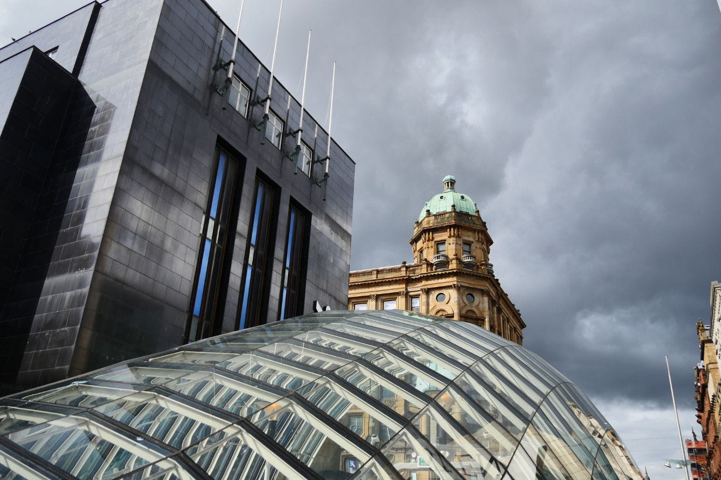 A glass-domed entrance to a modern building contrasts with a historic stone dome under a cloudy sky in Glasgow, Scotland