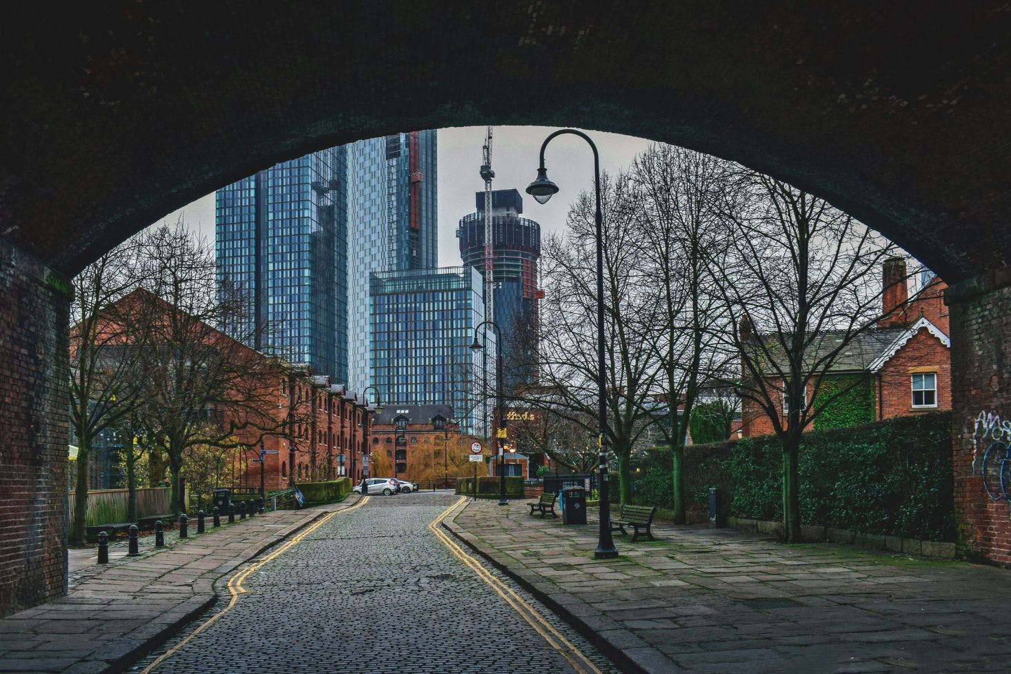 Street view under an arch with cobblestones, trees, and lampposts, leading to modern high-rise buildings in the background