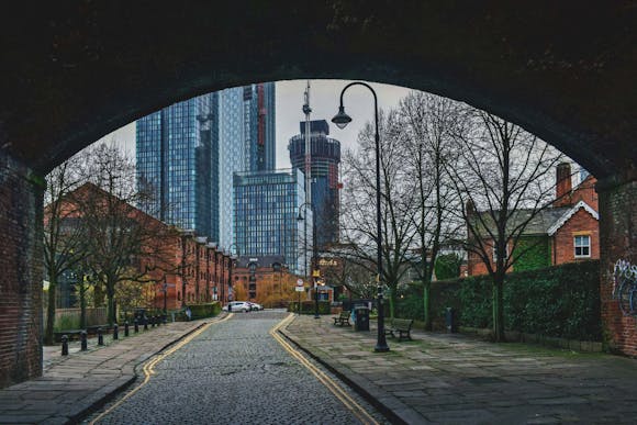 Street view under an arch with cobblestones, trees, and lampposts, leading to modern high-rise buildings in the background