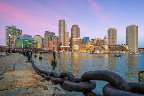 Boston cityscape at pink sunset, seen from the asphalt dock with a harbor chain along the waterfront