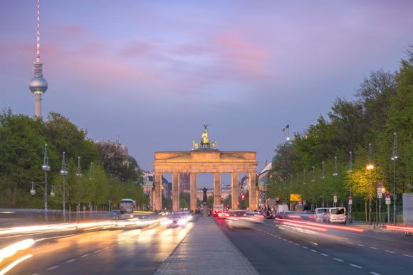 View of Berlin's Brandenburg Gate and TV Tower at sunset, with neon streaks of cars along Unter den Linden boulevard