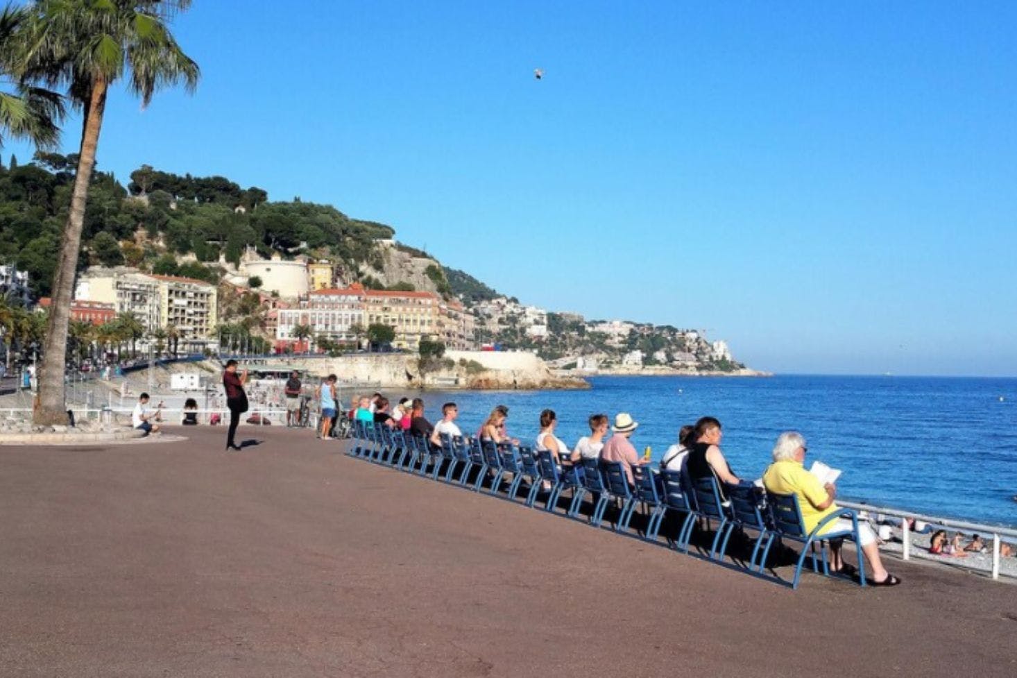 View of the Promenade des Anglais in Nice, with benches, tourists, palm trees, and the city in the background