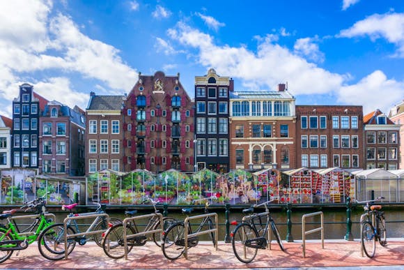 Bikes parked on a red cobblestone path in Amsterdam, with colorful brick houses, flower shops, and a canal in view