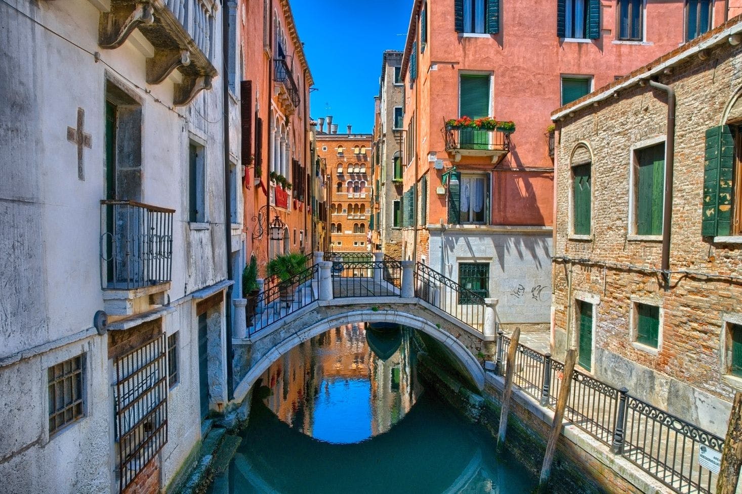 A narrow canal in Venice with colorful buildings, a small arched bridge, and a bright blue sky reflecting in the water.