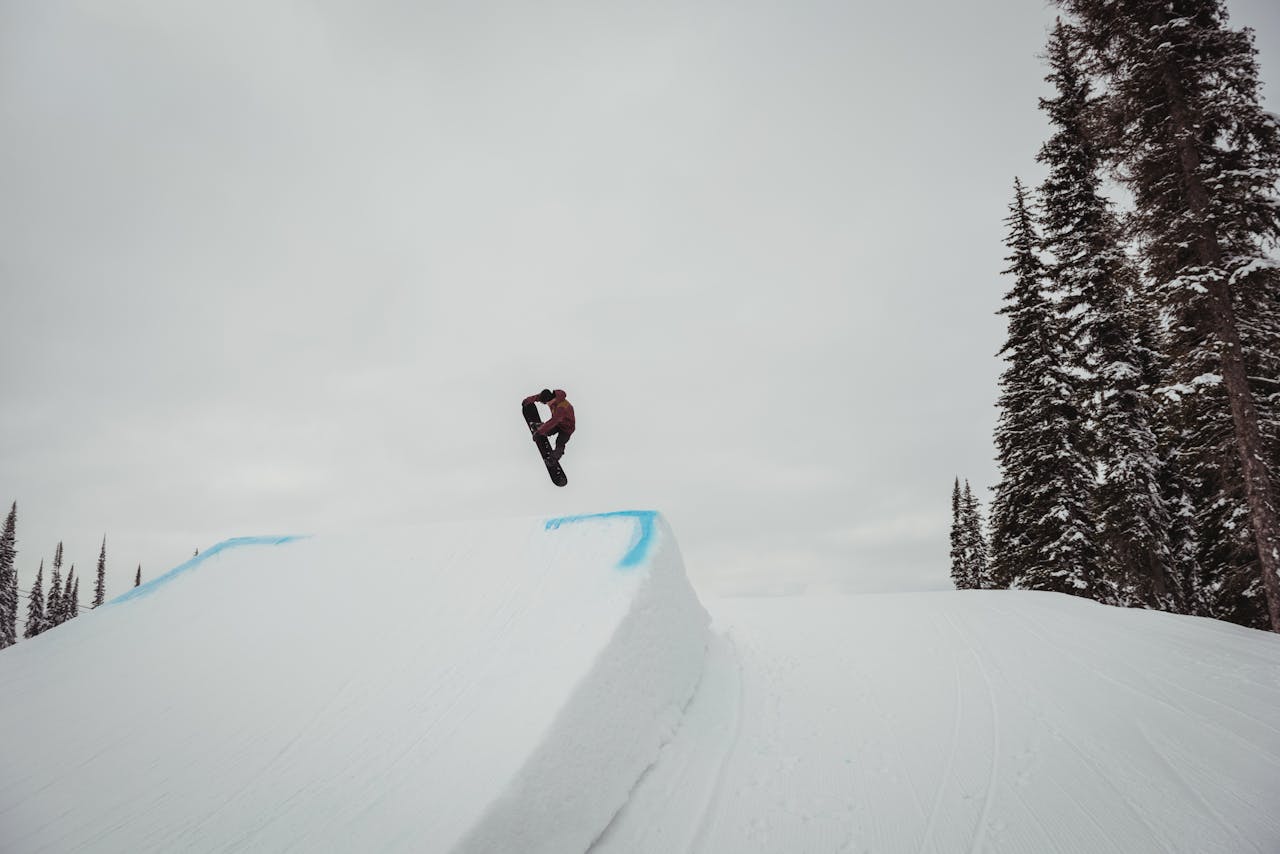 Snowboarder mid-air performing a trick on a snowy slope, surrounded by tall trees,