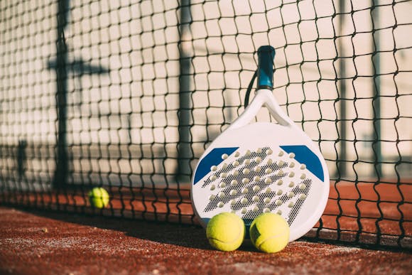 Padel racket and balls resting against a net on a clay court