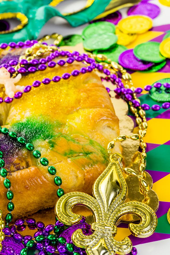 Close-up of a traditional King Cake surrounded by vibrant Mardi Gras beads, coins, and a gold fleur-de-lis