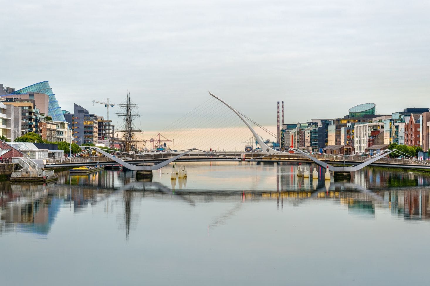 A view of Dublin’s Samuel Beckett Bridge reflected in the River Liffey with modern buildings and a tall ship line the riverbanks 