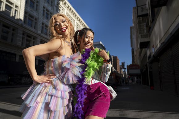 wo individuals dressed in vibrant and colorful outfits, smiling and posing on a sunny city street, celebrating the Sydney Gay and Lesbian Mardi Gras