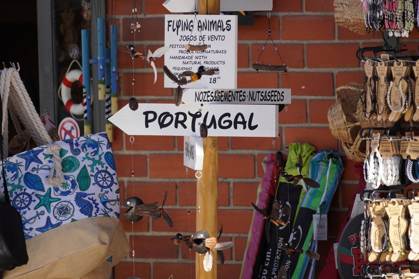 Street stall with colorful beach bags, bracelets, and trinkets, featuring an arrow sign reading "Portugal" in bold letters