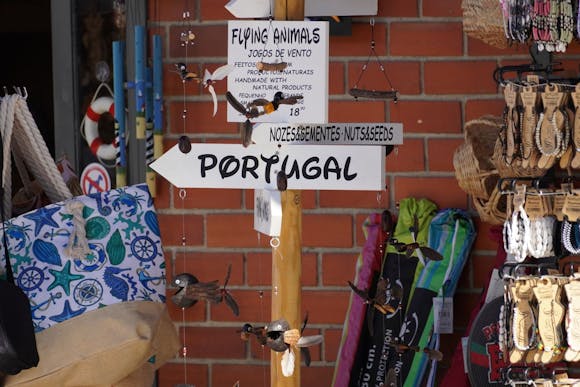 Street stall with colorful beach bags, bracelets, and trinkets, featuring an arrow sign reading "Portugal" in bold letters