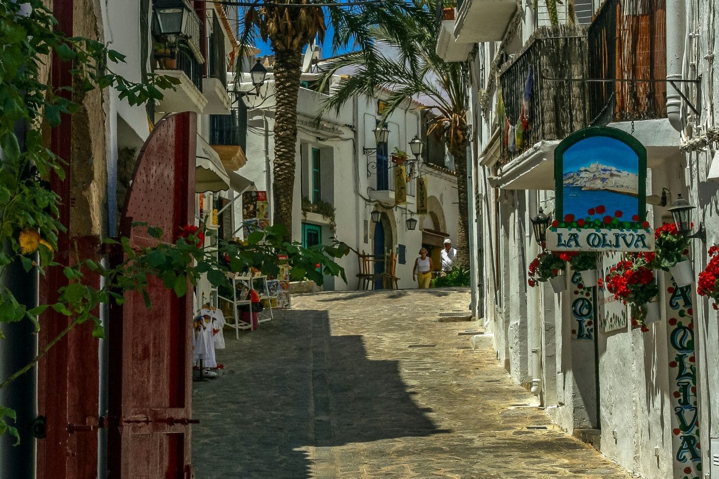 Cobbled street in Ibiza with old buildings, shops, balconies, and palm trees on a hilly path