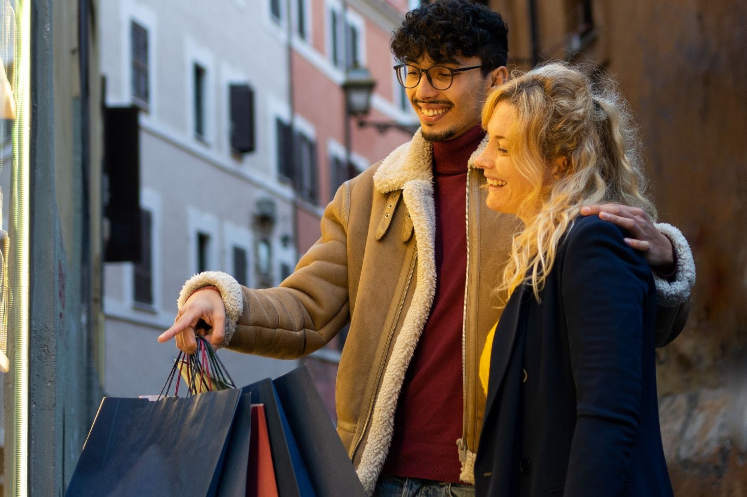 Curly-haired man with glasses and blonde woman smiling while holding shopping bags and window shopping together