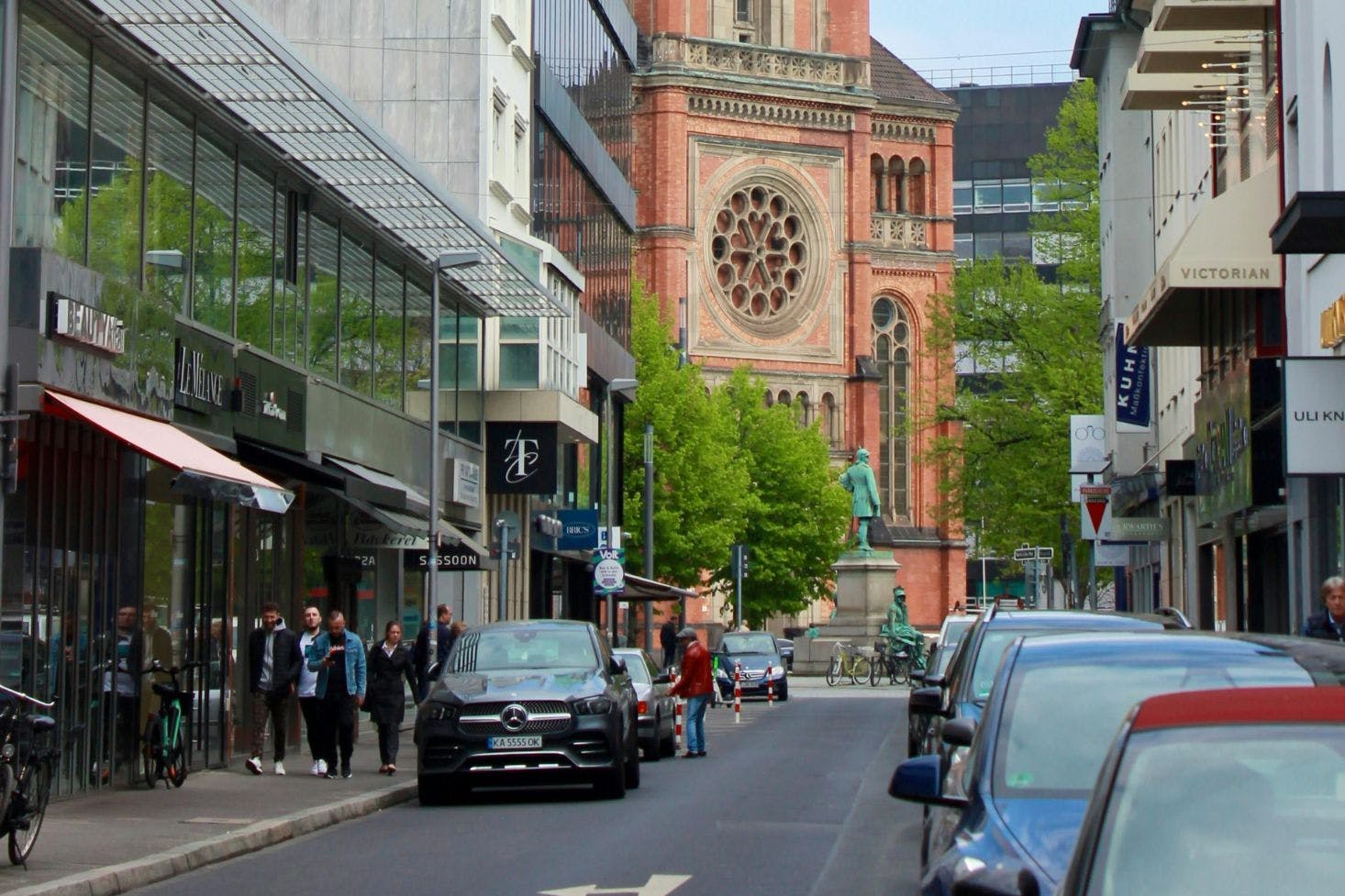 A shopping street in Düsseldorf with modern stores, a historic church, parked cars, and people strolling