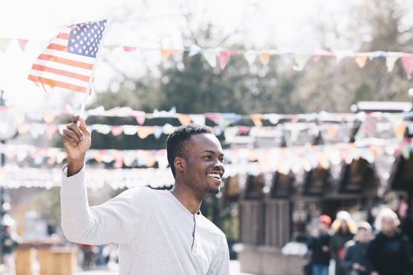 Smiling man waving an American flag at an outdoor celebration, surrounded by colorful bunting and a festive crowd