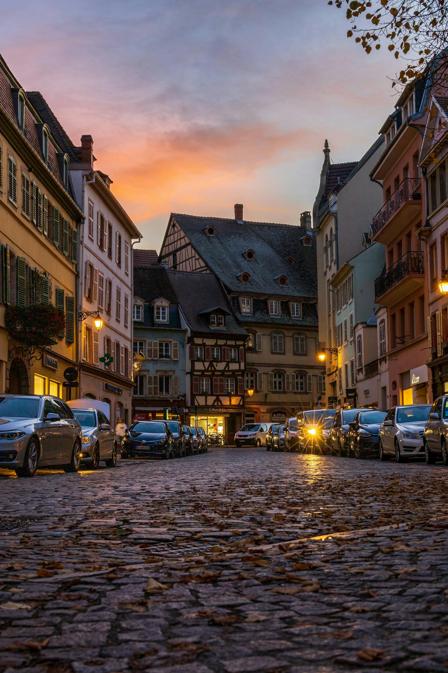 A shopping street in Colmar at sunset with cars parked along the road