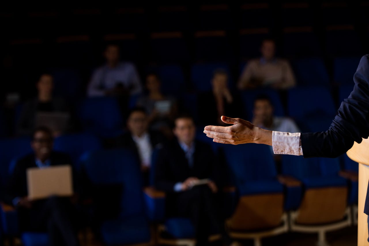 A business professional delivering a speech or presentation in a dimly lit conference room, with an audience seated in the background