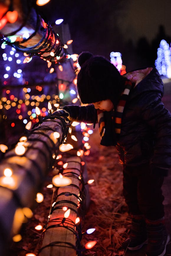 Young child dressed warmly in a winter jacket, hat, and scarf, leaning on a wooden fence wrapped in colorful, glowing string lights