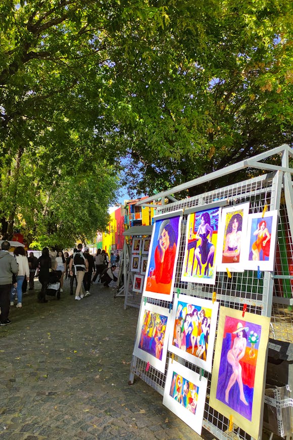 Colorful artwork displayed on a wire stand under lush green trees as visitors stroll through an art festival