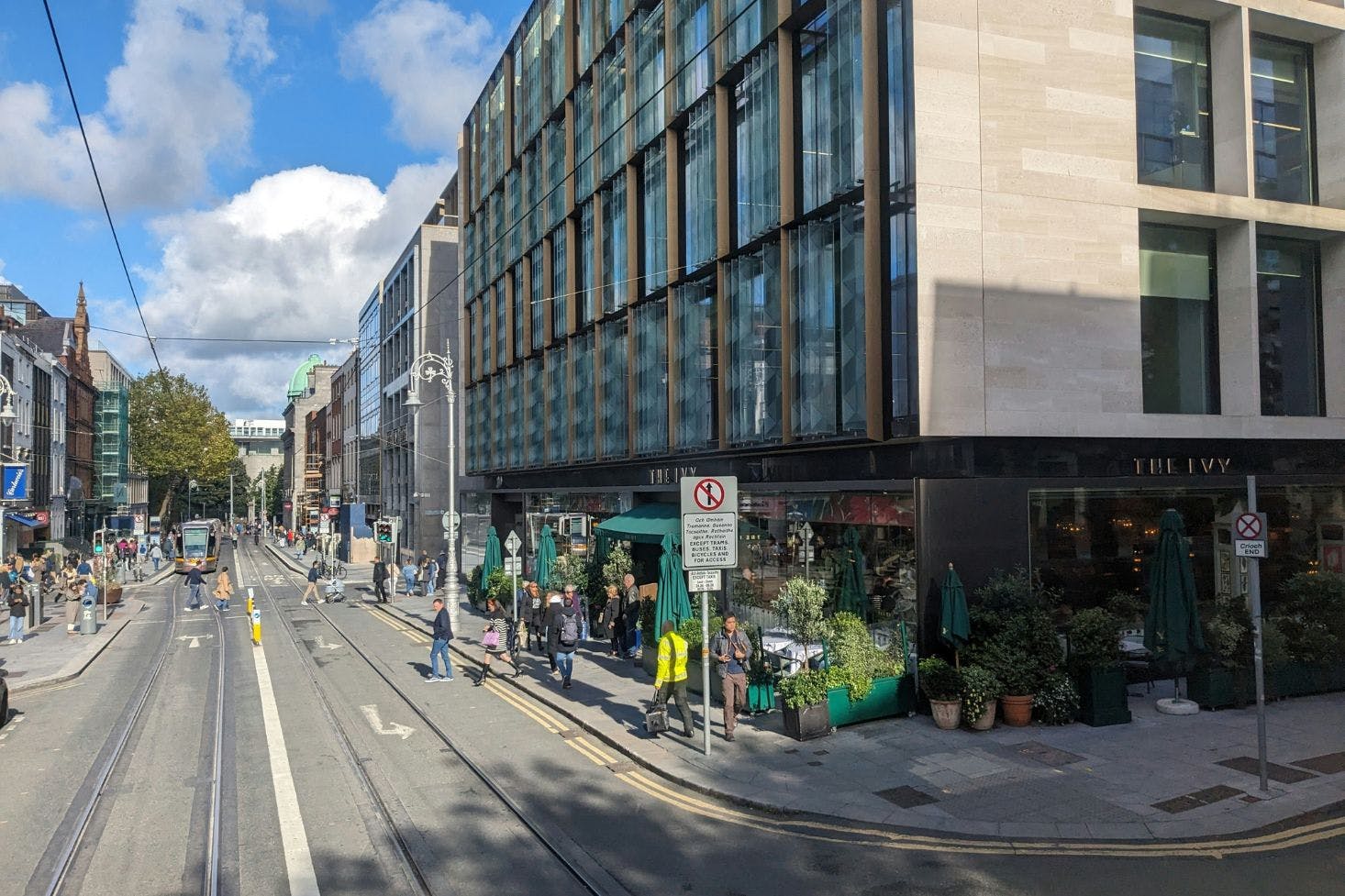 A street view of modern and historic buildings, a tram, people walking, and outdoor seating at The Ivy, Dublin