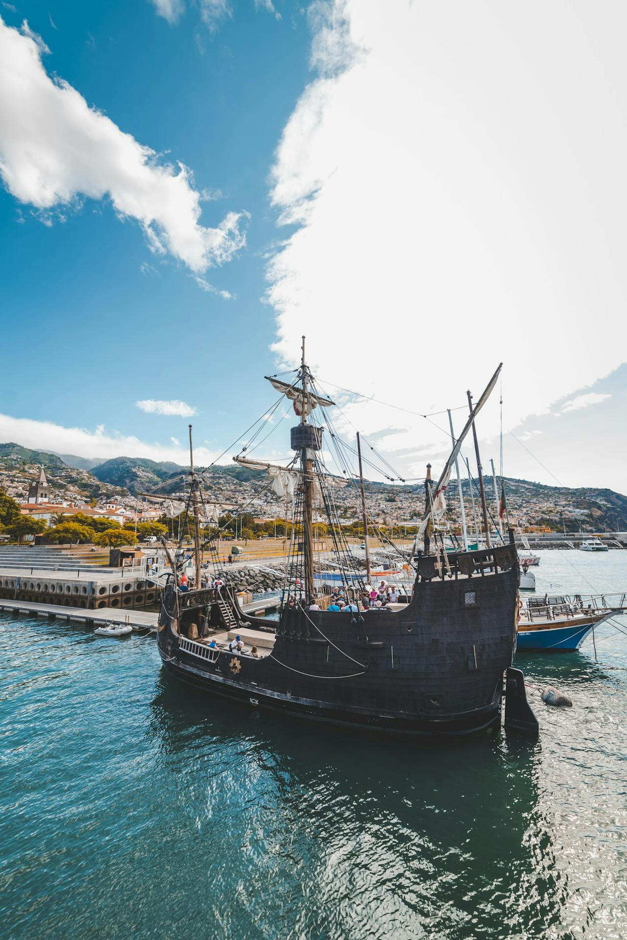 A historic wooden pirate ship docked at the harbor with stunning hills in the background