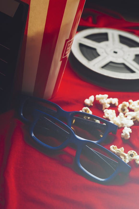 Two pairs of 3D glasses, scattered popcorn, and a vintage film reel on a red backdrop