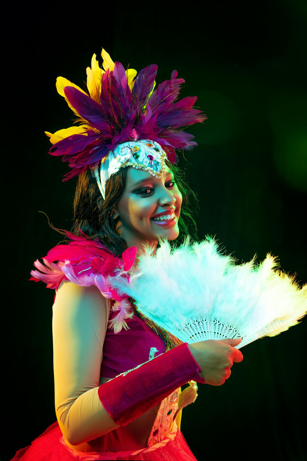 A vibrant carnival performer adorned in a colorful feathered headdress and sequined costume, smiling warmly while holding a delicate white fan