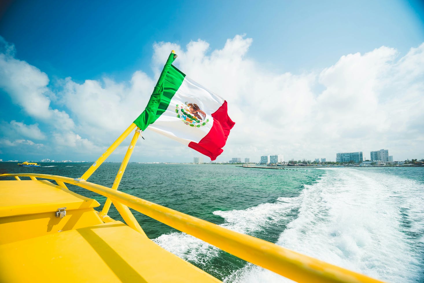 The rear of a yellow boat flying the Mexican flag leaving Cancun in its wake