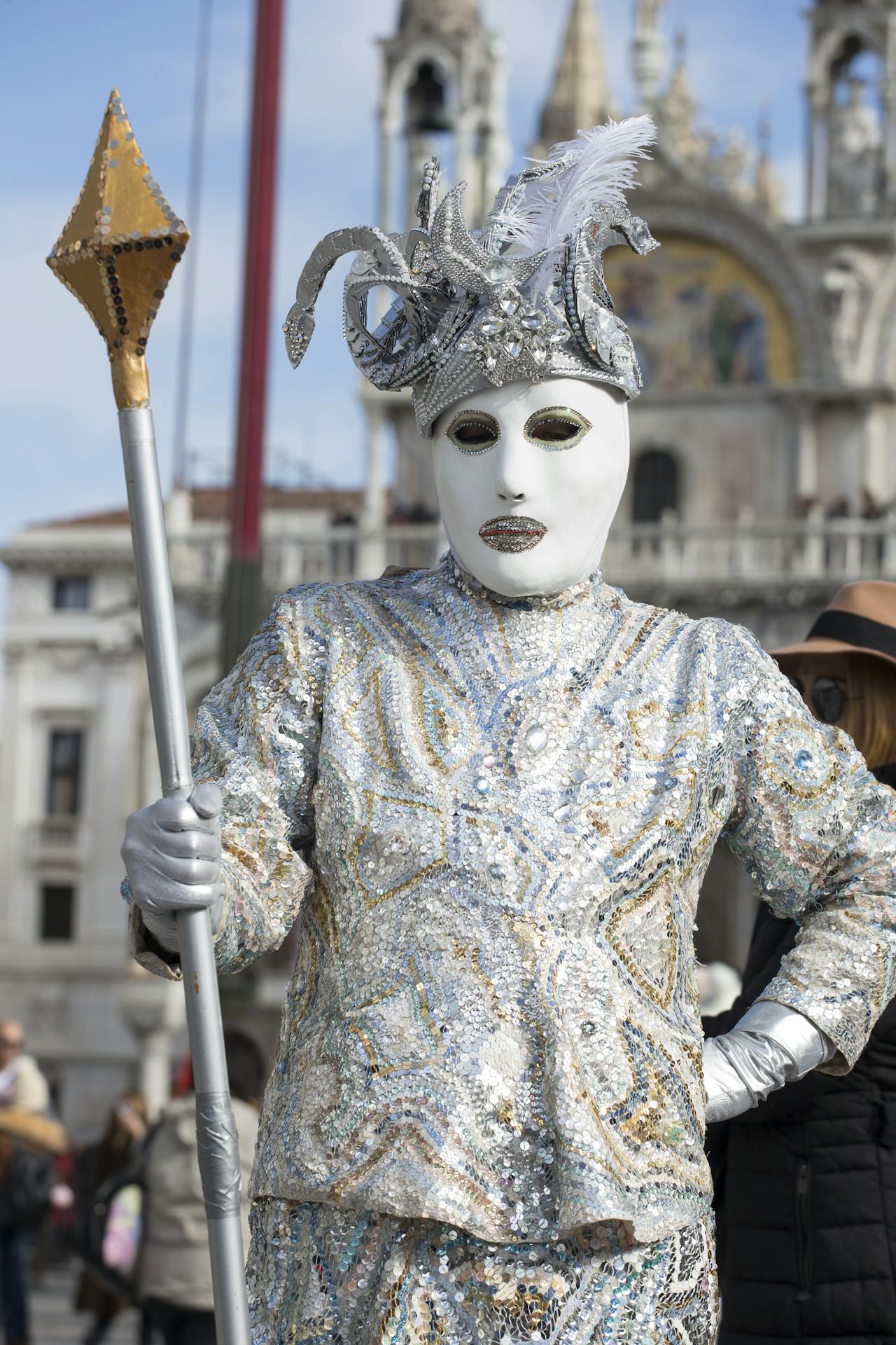 A person in a silver sequined costume and Venetian mask holding a staff during a carnival celebration.