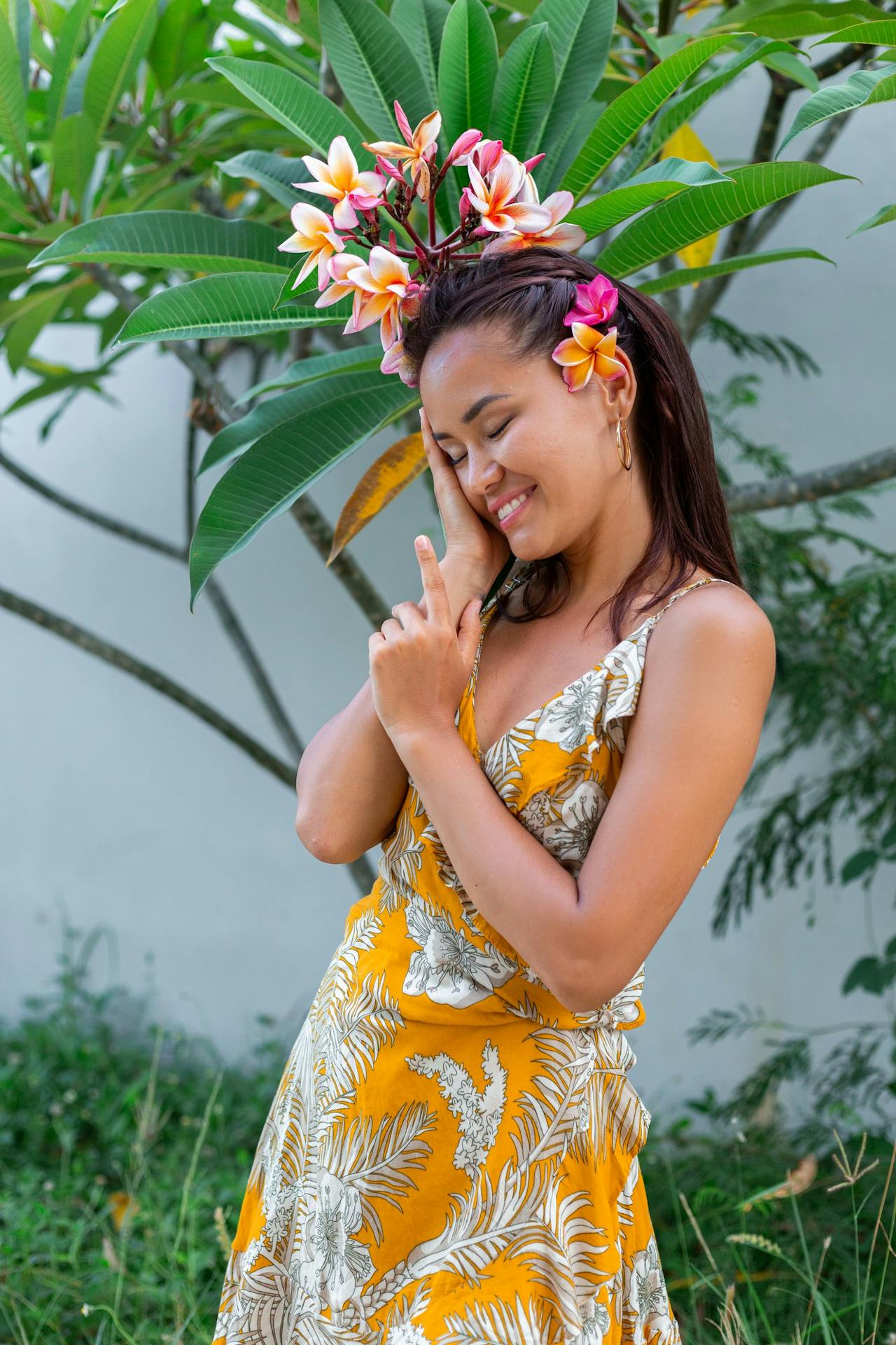 A woman wearing a yellow floral dress adorned with plumeria flowers in her hair smiles while standing outdoors