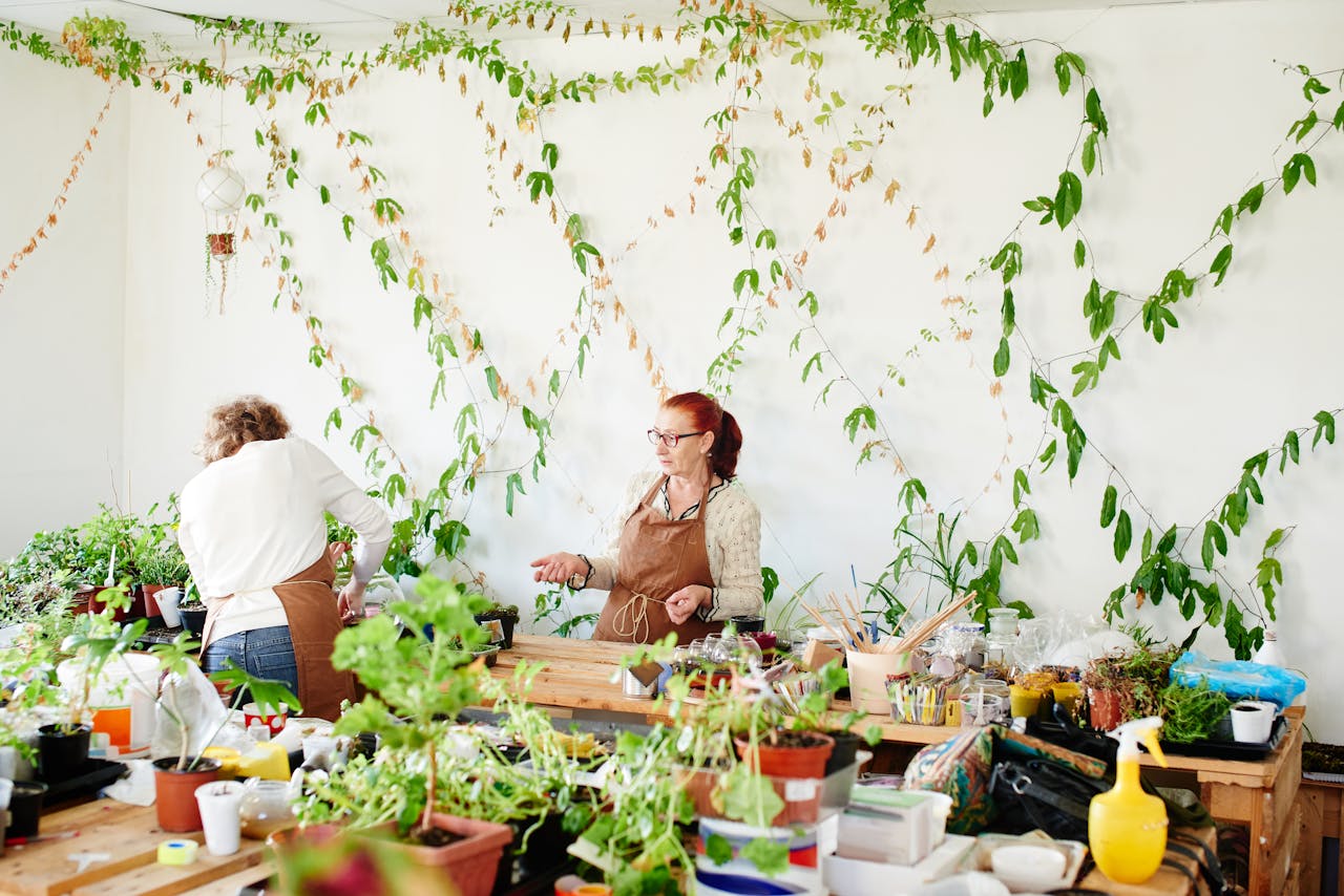 Two women wearing aprons tending to a variety of potted plants in a bright, greenery-filled workshop