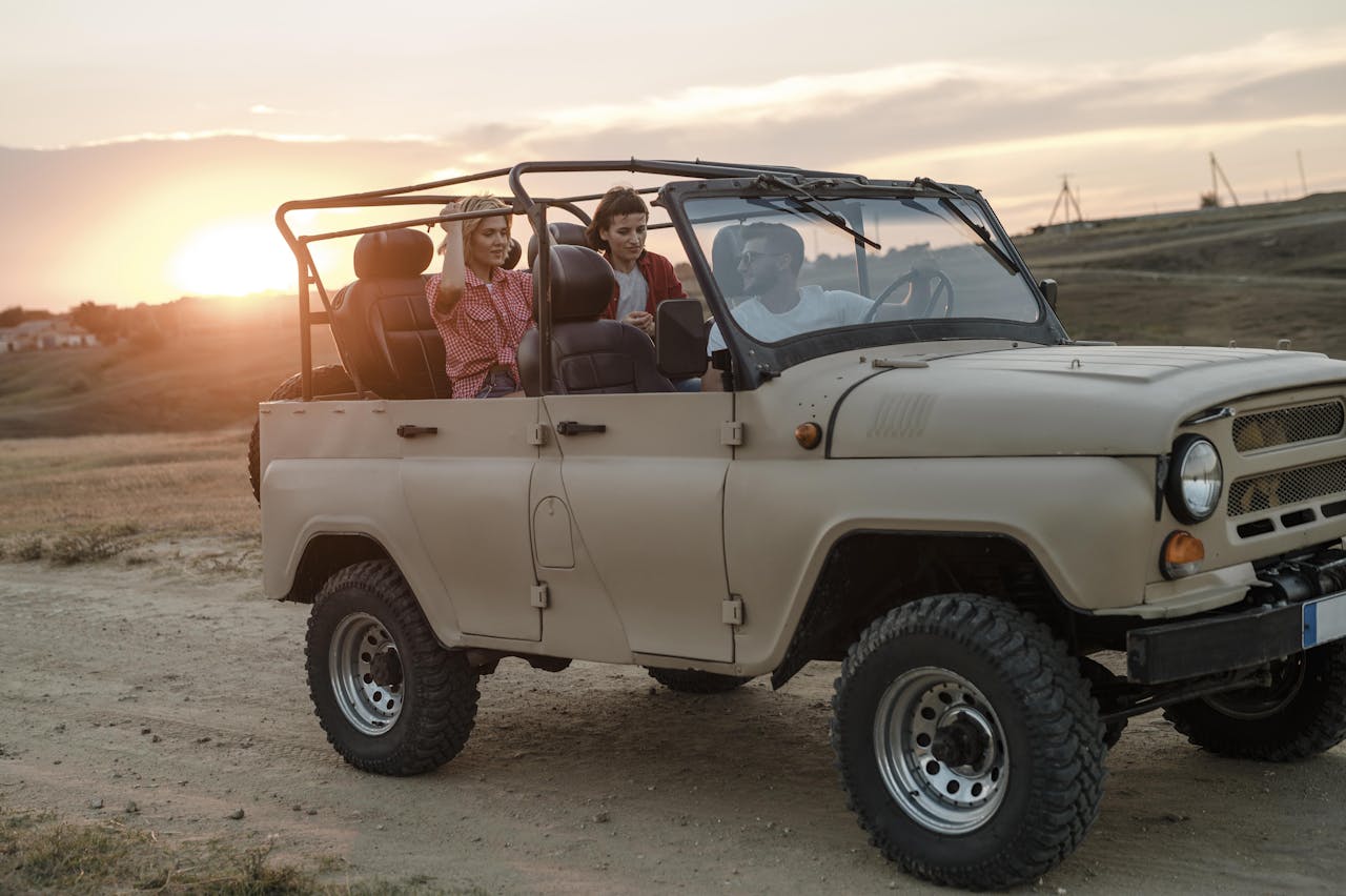 A rugged, tan off-road Jeep with large tires and an open-top design, parked on a dirt trail at sunset