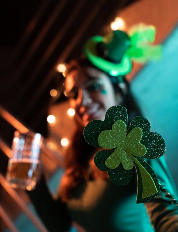 Festive woman in green with a glittery shamrock, mini hat, and beer mug, celebrating St. Patrick's Day amid warm string lights