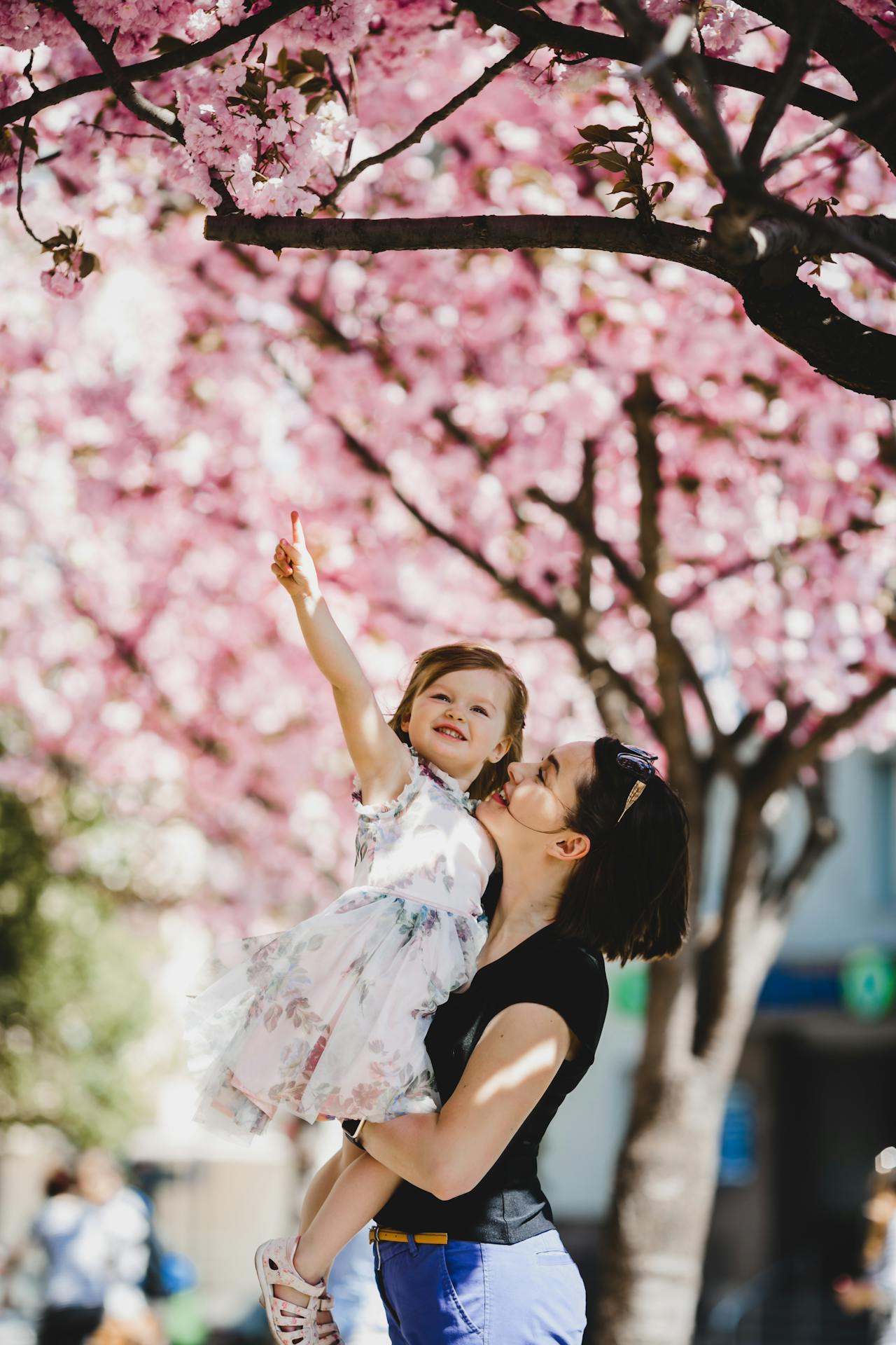 Mom standing and holding her smiling daughter under cherry blossom trees