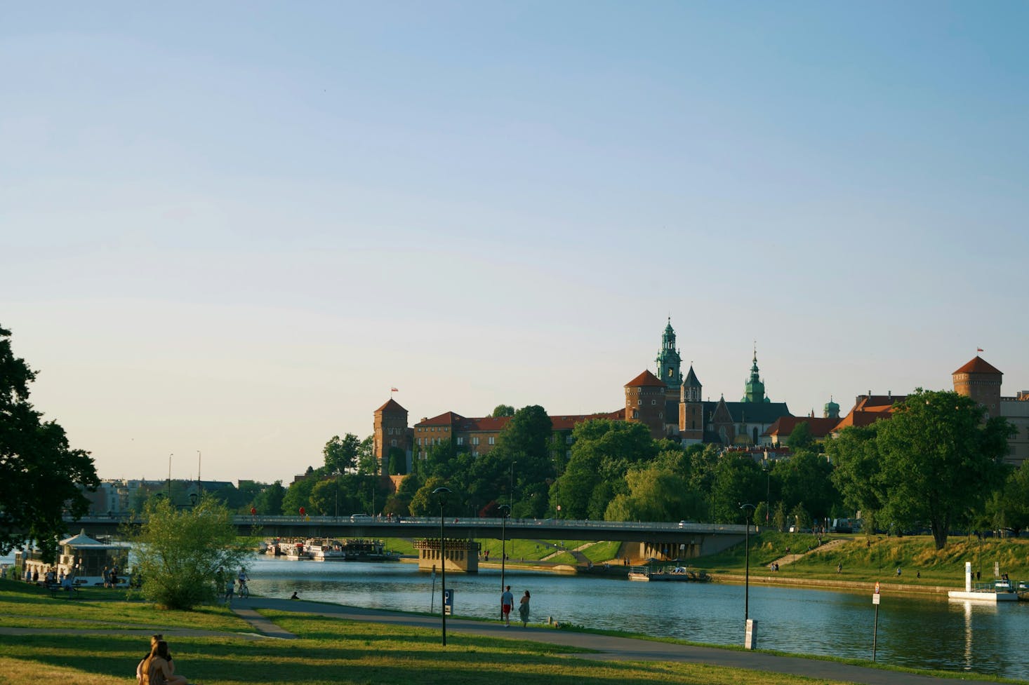 Large historic building overlooking a river and park, with a bridge in the center