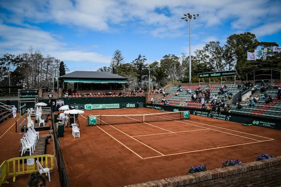 An empty red clay tennis court during a warm sunny day outdoors, with spectators slowly taking their seats