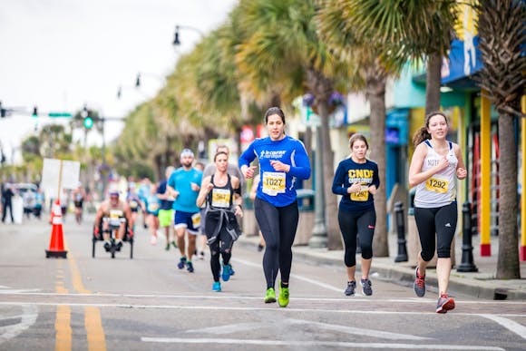 Runners wearing bib numbers running through a street lined with Palm trees during a marathon