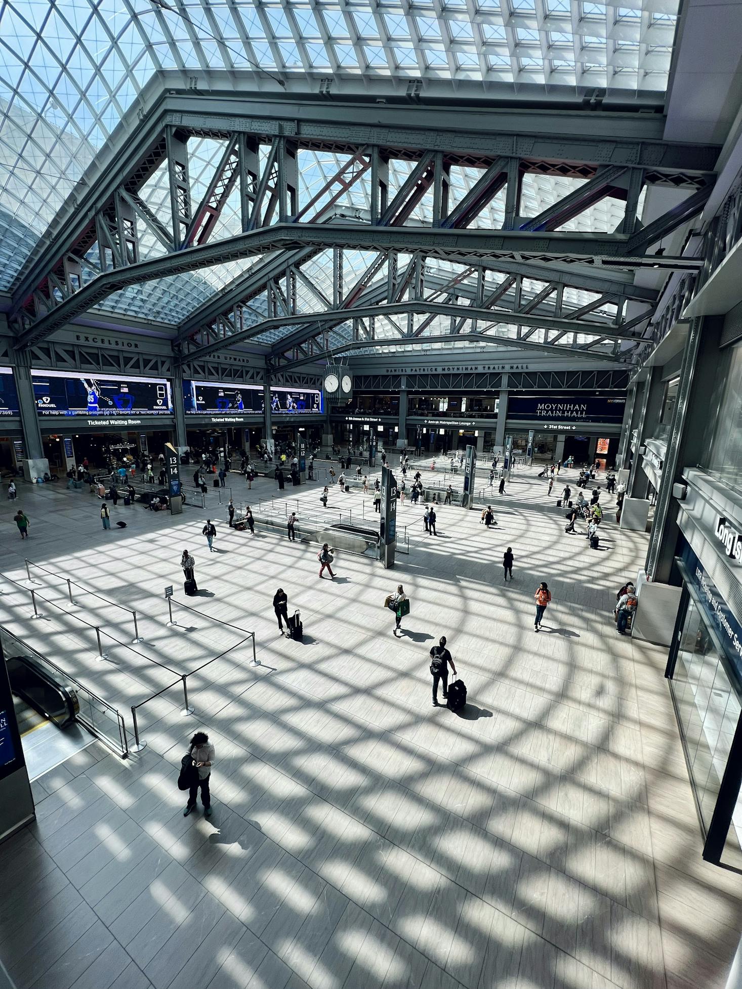 Natural light filters through the ceiling, creating a pattern of shadows at the bustling Moynihan Train Hall.