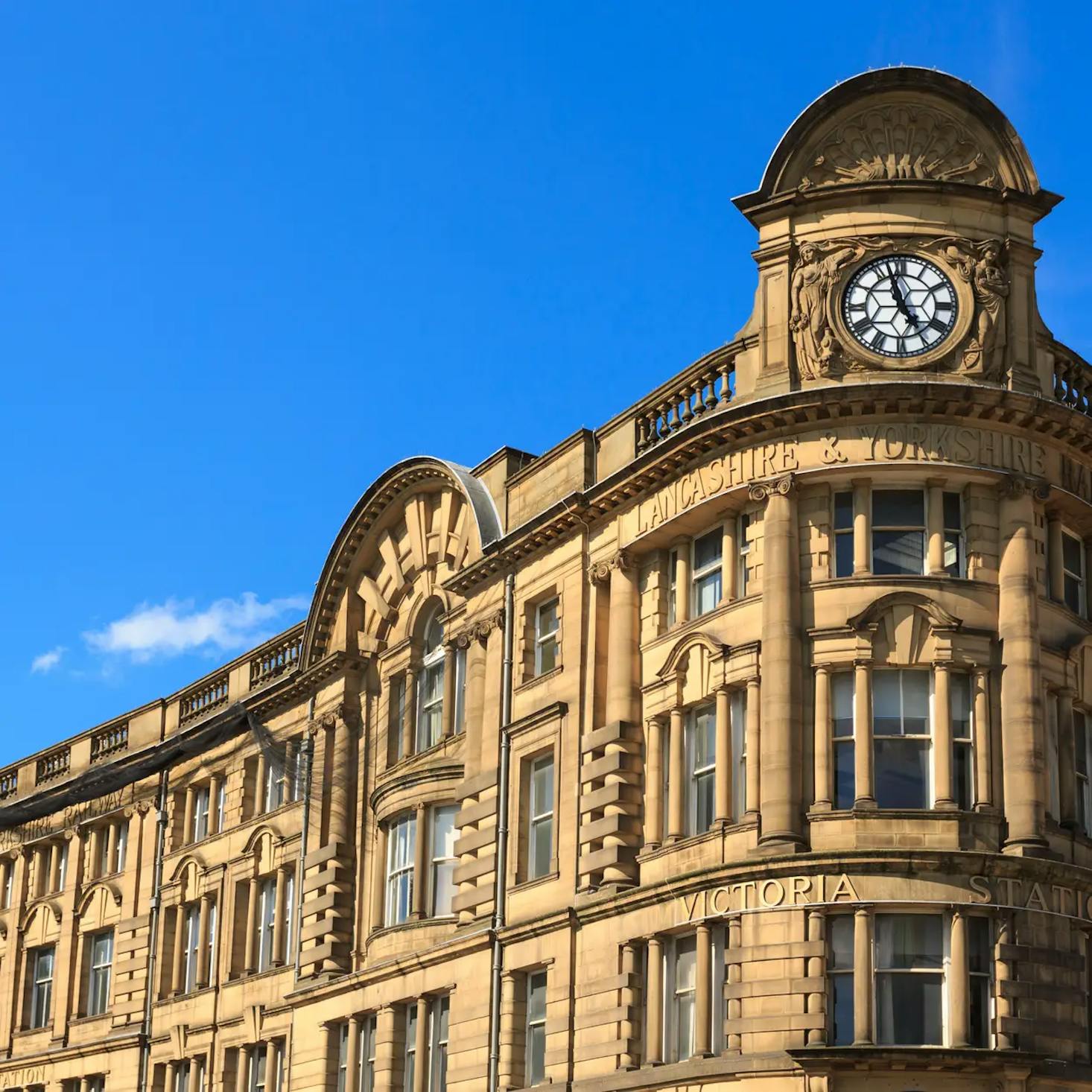 The Manchester Victoria Station, featuring a clock and intricate architectural details against a clear blue sky.