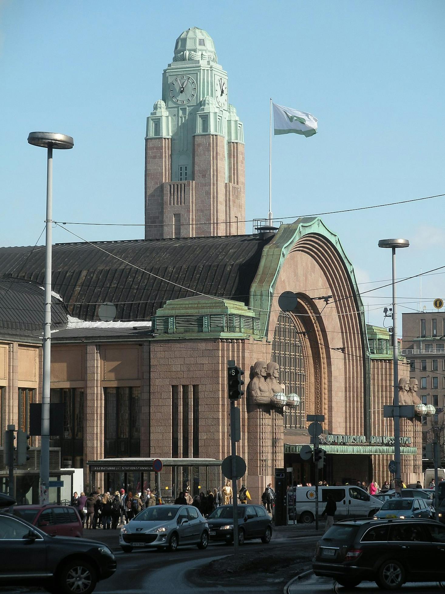 The exterior of Helsinki Central Station, featuring its iconic clock tower, art deco architecture, and a bustling street with cars and pedestrians in the foreground.