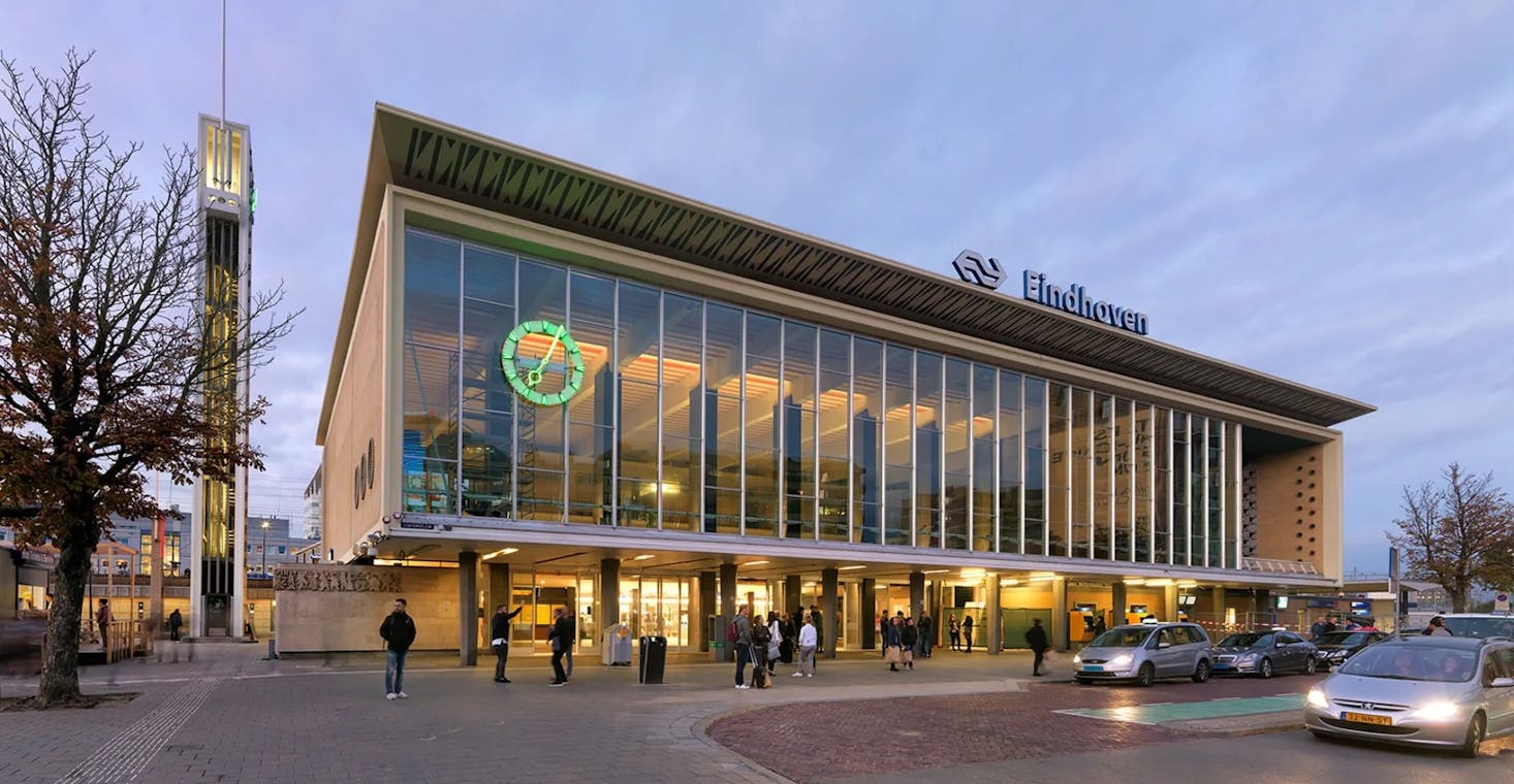 The front facade of Eindhoven train station in the evening, with people walking and cars parked in front.