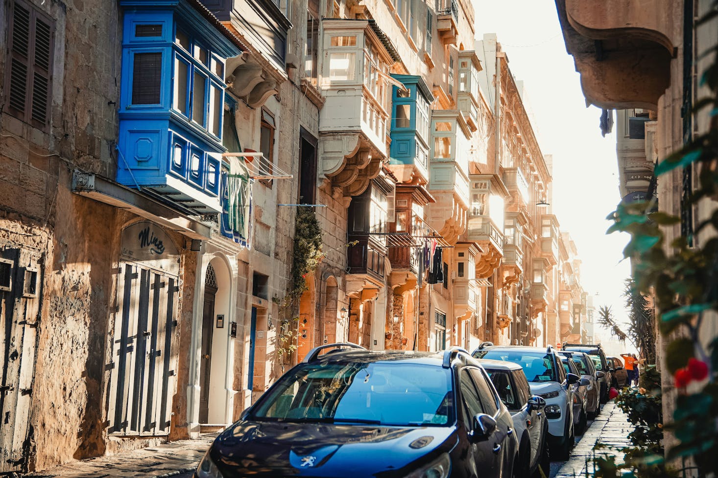 A narrow street lined with parked cars and historic buildings featuring colorful, enclosed balconies in Valletta.