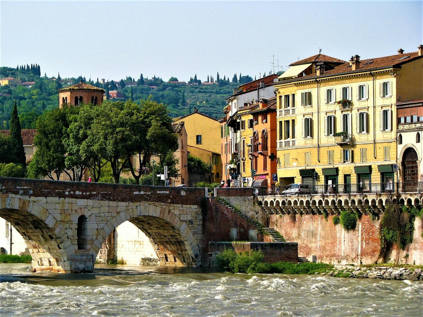 Scenic view of a river with a stone bridge, historic buildings, and lush greenery in the background in Verona