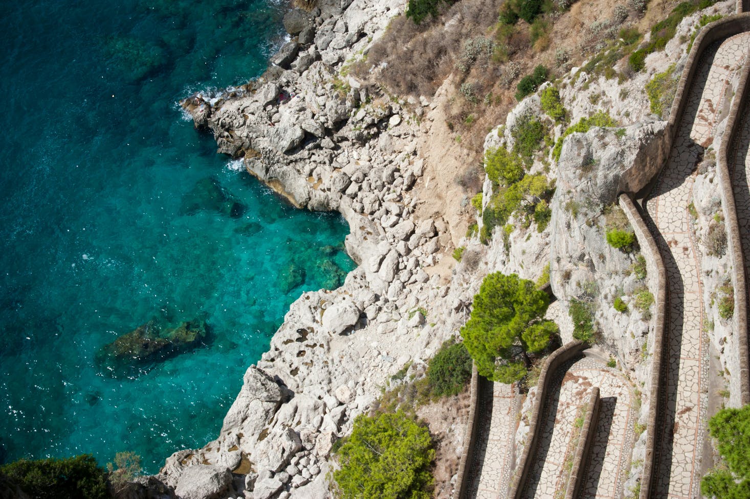 Vista dall'alto sul mare di Capri, con acqua cristallina e stradina acciottolata a serpente che scende verso la spiaggia