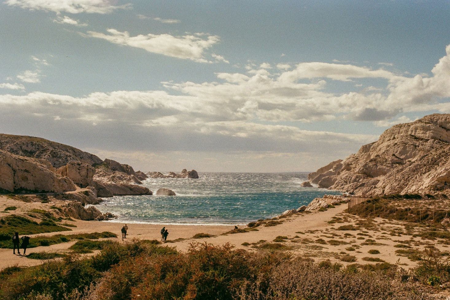 One of the sandy beaches near Marseille with patches of grass, rocks, and sea with people exploring it