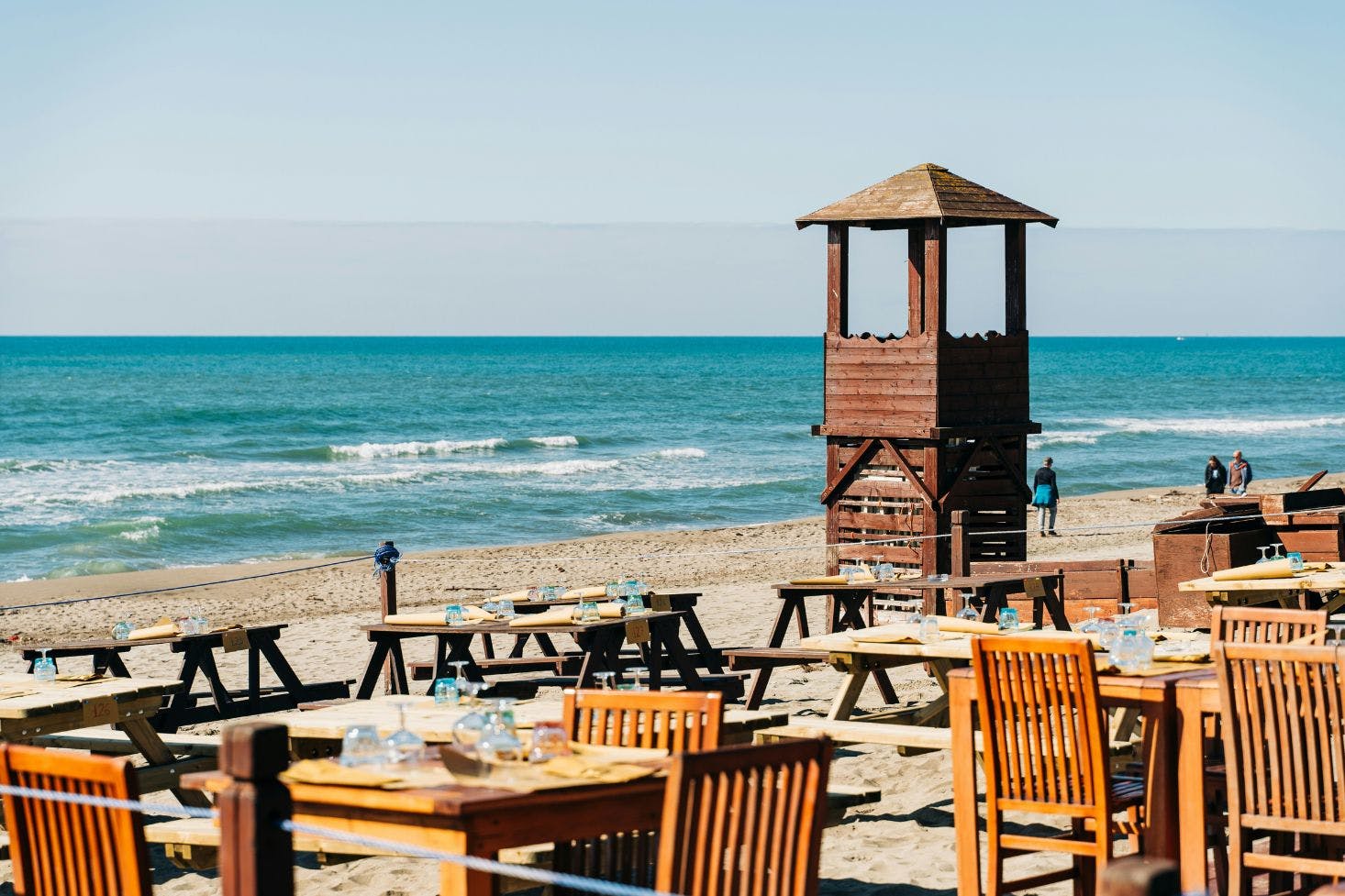 Wooden tables and chairs on a sandy beach near Rome, with a lifeguard tower and blue sea in the background