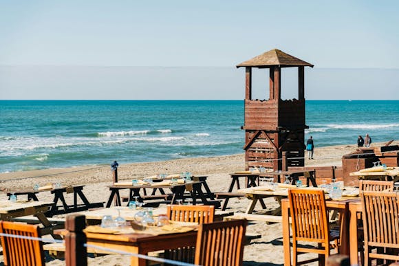 Wooden tables and chairs on a sandy beach near Rome, with a lifeguard tower and blue sea in the background