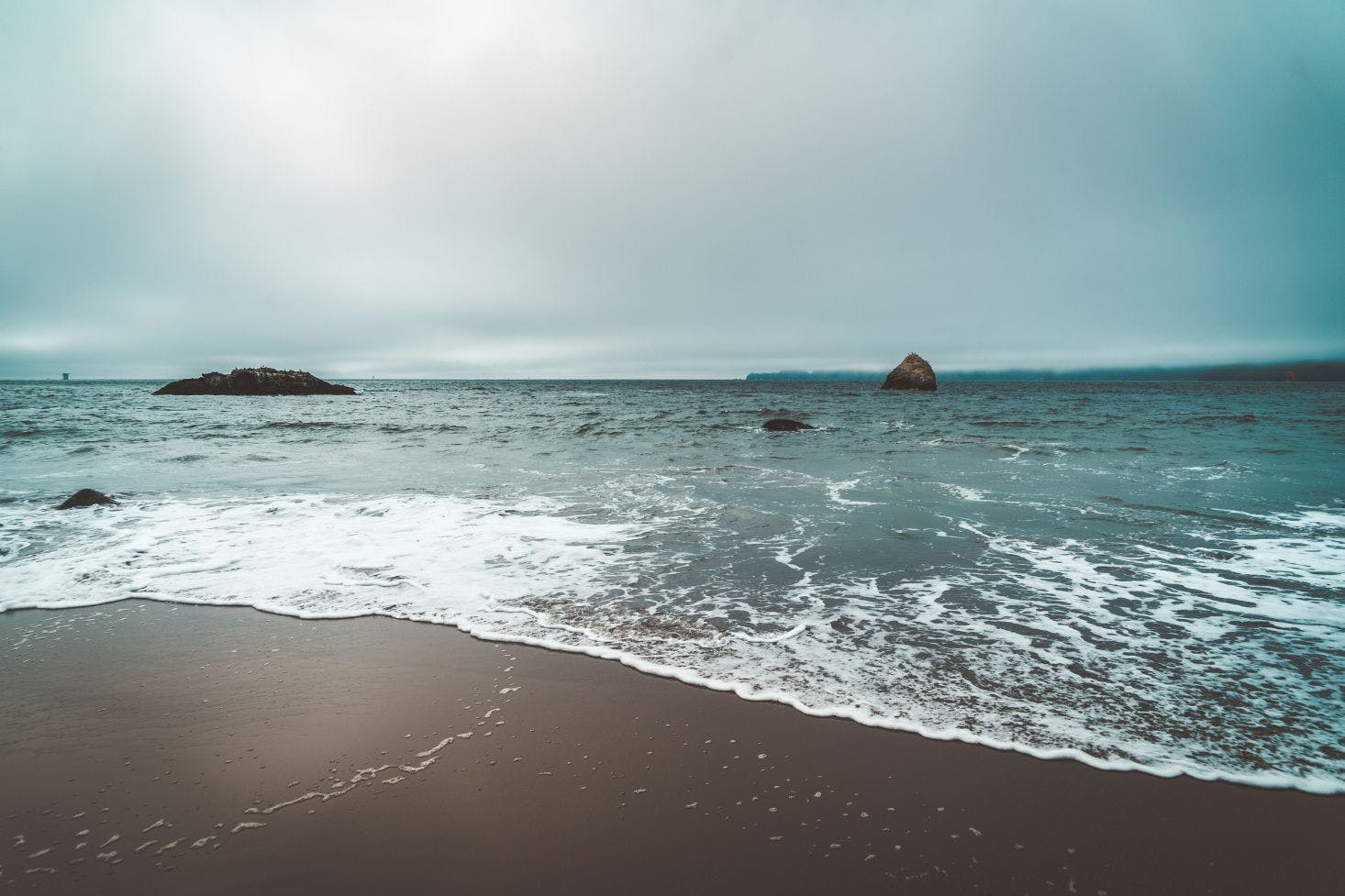 Dark brown sandy beach in San Francisco with foamy waves rolling in under a dark gray sky.