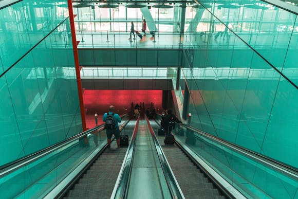 Passengers with luggage descend the escalator in a glass-wall Rome airport terminal