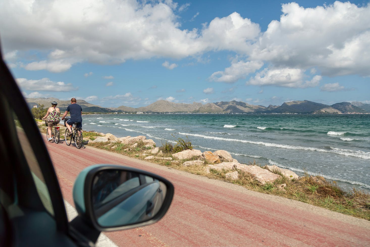 Vista da un'auto di mare mosso e montagne sullo sfondo, con due turisti in bici sulla strada lungomare